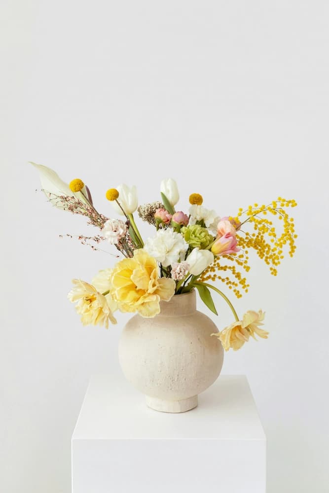 Arranjo moderno com flores brancas e amarelas no vaso branco em cima de um pedestal.